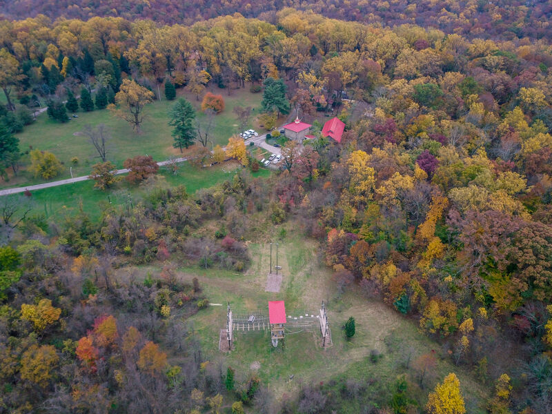 The image shows an aerial view of a landscape with a mix of trees, grass, and buildings. The trees are in various stages of color change, indicating it is likely autumn. There are a few buildings visible, including what appears to be a house or lodge with a red roof. There is also a structure that might be a shooting range.